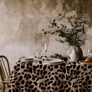 Puede incluir: Un mantel de mesa de estampado de leopardo marrón y negro con un jarrón de cerámica blanco lleno de ramas secas y un candelero blanco con una vela encendida. Hay dos platos blancos, dos copas de vino y una bandeja de madera con panecillos en la mesa.