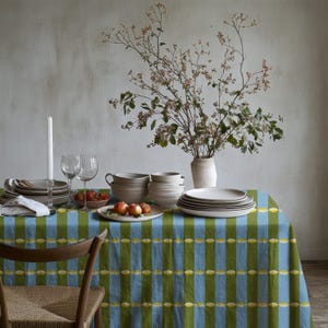 May include: A dining table covered in a blue and olive green striped tablecloth with small yellow oval accents. The table is set with stacks of white ceramic plates and bowls, two clear glass wine glasses, a white candle, and a bowl of apples.