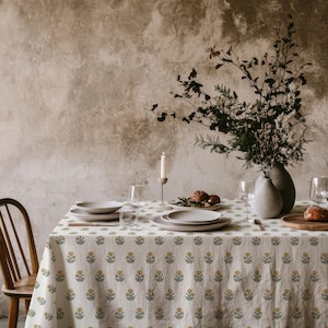 Puede incluir: Una mesa de comedor preparada para una comida, con un mantel crema con un estampado floral amarillo y verde. La mesa está puesta con platos, vasos y un jarrón con flores. También se ven una vela encendida y una silla de madera.