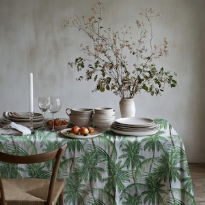 Peut inclure: Une nappe blanche à imprimé palmier vert présente une table dressée avec des bols, des assiettes et des verres en céramique blanche. Un vase avec des branches séchées se trouve au centre de la table.