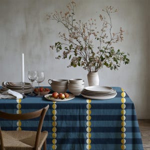 May include: A blue and dark blue striped tablecloth with vertical yellow circular patterns, set with a stack of ceramic bowls, plates, wine glasses, a white candle, and a vase containing branches, alongside a bowl of small red fruits.