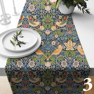 May include: A table runner featuring a repeating pattern of birds, strawberries, and floral motifs in shades of navy blue, green, orange, and cream. The design includes stylized foliage and birds perched among flowers on a dark blue background.