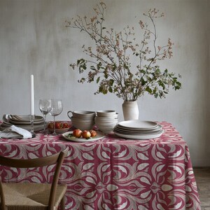 May include: A table covered with a red and beige floral patterned tablecloth, featuring a stack of white ceramic plates, bowls, two clear wine glasses, a tall white candle, a small bowl of red berries, and a vase with leafy branches.