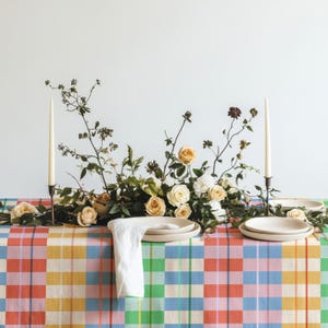 Könnte beinhalten: Ein gedeckter Tisch mit einer karierten Tischdecke in Blau, Gelb, Rosa und Grün. Ein Blumenarrangement aus cremefarbenen Rosen und Grünpflanzen steht zwischen zwei hohen weißen Stabkerzen. Zwei weiße Keramikteller und eine weiße Serviette liegen bereit.