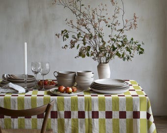 Peut inclure: Une nappe rectangulaire ornée d'un motif à carreaux répétitif composé de rectangles vert citron et de carrés marron foncé. La table est dressée avec des assiettes et bols en céramique blanche, deux verres à vin, une bougie et un vase de branches.
