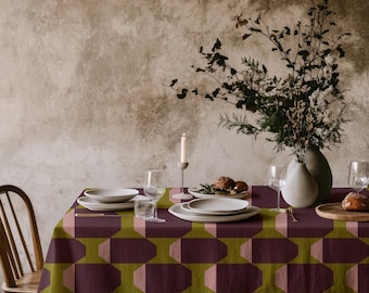 Könnte beinhalten: Eine rechteckige Tischdecke mit einem geometrischen Muster aus dunkelvioletten und olivgrünen Achtecken. Der Tisch ist mit weißen Keramiktellern, klaren Weingläsern, einer weißen Kerze im Halter und einer Vase mit getrockneten Zweigen gedeckt.