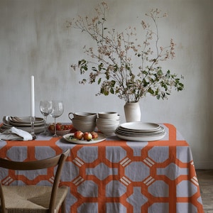 May include: A dining table set with a gray tablecloth featuring an orange geometric design. The table is set with plates, bowls, glasses, and a vase of branches. Apples and a lit candle are also present.