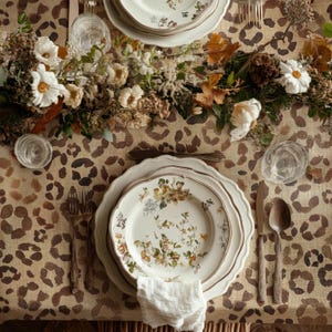 May include: A table setting with floral patterned plates, silverware, and glasses. The plates have a scalloped edge and a gold rim. The tablecloth has a brown and tan animal print. A floral arrangement is in the center.