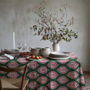 May include: A dark green tablecloth featuring a repeating pink and white leaf pattern. The table is set with stacks of white ceramic bowls and plates, two clear glass wine glasses, a white taper candle in a holder, and a small bowl of apples.