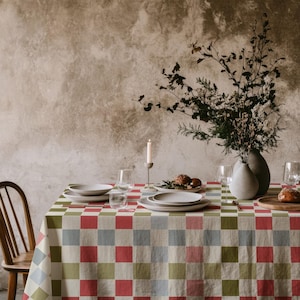May include: A rectangular tablecloth featuring a repeating checkered pattern in muted shades of red, olive green, and light blue. The fabric covers a dining table set with white ceramic plates, clear glass stemware, a candle, and a vase.