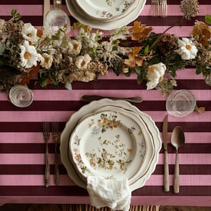 May include: A dining table set with floral plates, cutlery, and a white napkin. The plates are arranged on a pink and burgundy striped tablecloth. A floral centrepiece with white flowers and greenery adorns the table.