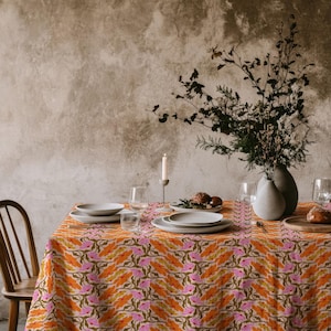 May include: A rectangular tablecloth featuring a repeating floral pattern with orange and pink petals on a light background, draped over a dining table set with white ceramic plates, clear glassware, a candle holder, and a vase with dried branches.
