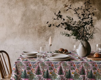 May include: A rectangular tablecloth featuring a repeating pattern of dark green pine trees, colorful floral clusters, and leafy branches on a light pink background. The fabric drapes over a table set with white ceramic plates, glassware, and a candle.
