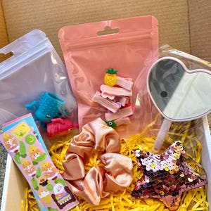 May include: A gift box filled with various accessories. Includes pink hair clips with fruit designs, a pink sequined star keychain, a heart-shaped mirror, a pink scrunchie, and stickers. The box is lined with yellow shredded paper.