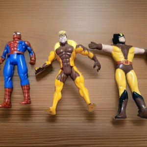 May include: Three plastic superhero action figures standing in a row. The left figure is red and blue with a web pattern. The center figure is yellow and brown with blonde hair. The right figure is yellow and brown with a black mask and extended arms.
