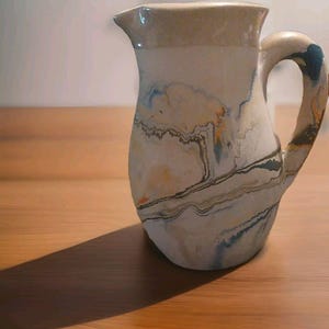 May include: A ceramic pitcher with a marbled design in shades of white, beige, blue, and orange. The pitcher has a handle and a spout. The pitcher is sitting on a wooden surface.