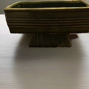 May include: A green ceramic planter with a rectangular top and a square base. The planter has a textured surface and a raised lip around the top.