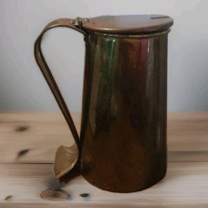 May include: A vintage copper-colored metal pitcher with a hinged lid and a curved handle. The pitcher has a cylindrical body and sits on a wooden surface. The handle is attached to the side of the pitcher.