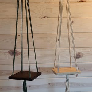 May include: Two macrame plant hangers. One has a dark brown square wooden shelf and olive green rope. The other has a light wood shelf and natural colored rope. Both are suspended from wooden rings.