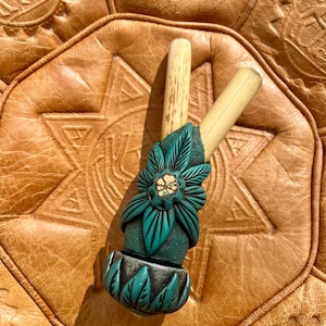 Peut inclure: Une pipe en bois sculpté de couleur vert sarcelle et marron avec un motif floral. La pipe est posée sur une surface en cuir marron.