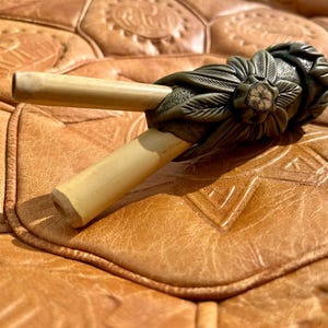 Peut inclure: Une pipe en bois sculptée de couleur verte et brune avec un motif floral décoratif. La pipe a deux tiges en bois et une petite pierre ronde blanche au centre du motif floral.