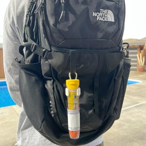Peut inclure: Un sac à dos noir avec le texte "The North Face" sur le devant. Un tube jaune et blanc avec une étiquette blanche est attaché au sac à dos. L'étiquette porte le texte "RECON".