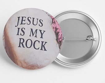 May include: A round button with the text "JESUS IS MY ROCK" in black letters. The button has a light-colored background with a textured appearance and a pink floral accent. The back of the button is silver.
