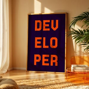 May include: A framed art print with the word "DEVELOPER" in orange pixelated letters against a dark blue background. The artwork is displayed in a room with natural light, a plant, and a modern orange side table.