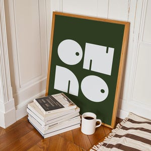 Op de afbeelding: Een ingelijste kunstprint met een donkergroene achtergrond en witte abstracte vormen. De print leunt tegen een muur naast een stapel boeken en een witte mok gevuld met koffie. Een gestreept tapijt is zichtbaar.