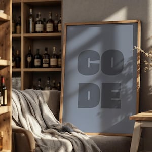 May include: A framed poster displaying the word "CODE" in large, grey letters. The poster leans against a chair draped with a grey blanket. A wooden cabinet with bottles is in the background. A vase of dried flowers sits on a small table.