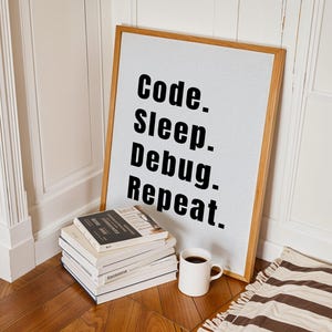 Peut inclure: Une impression encadr&eacute;e avec le texte "Code. Sleep. Debug. Repeat." sur un fond blanc. Le cadre est en bois et a une finition naturelle. L'impression est appuy&eacute;e contre un mur blanc, et il y a quelques livres empil&eacute;s sur un sol en bois devant elle.