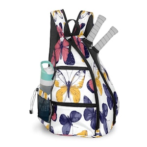 Puede incluir: Una mochila blanca con un estampado de mariposas en amarillo, rosa y azul. La mochila tiene una parte trasera de malla negra, un bolsillo lateral con una botella de agua y dos raquetas de tenis. El texto "WATER" es visible en el bolsillo lateral.