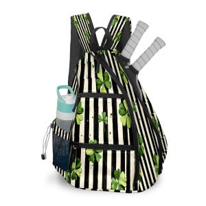May include: A black and white striped backpack with green shamrock accents. The backpack has a side pocket with a water bottle and two tennis rackets. The bag has black straps and a black zipper.