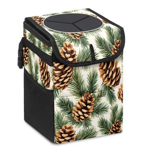 May include: A square, insulated car trash can with a black lid and side pocket. The exterior features a cream-colored background with a repeating pattern of brown pine cones and green pine needles. A black handle and trim complete the design.