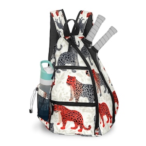 May include: A white backpack with a black mesh back and a zipper closure. The backpack features a repeating pattern of red and gray leopards. A water bottle and two tennis rackets are visible.