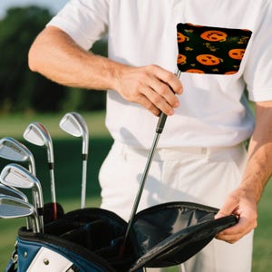 May include: A person holding a golf club with a black handle and a Halloween-themed head cover. The head cover has a black background with orange pumpkins and green leaves. A golf bag with several clubs is in the background.