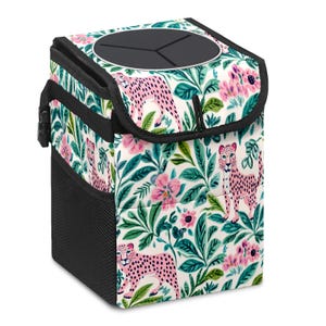 May include: A white, patterned car trash can featuring pink cheetahs and pink flowers amidst green foliage. The trash can has a black top with a circular opening and a black mesh side pocket. A black handle is attached to the side.
