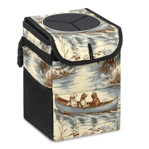May include: A beige and blue patterned car trash can featuring a black lid with a circular opening. The design depicts three dogs in a canoe on a river. The can has a black mesh side pocket and a black handle.