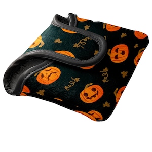 May include: A black Halloween-themed golf club head cover with a repeating pattern of orange jack-o'-lanterns and gold leaf accents. The cover has a gray trim and is designed to protect golf clubs.