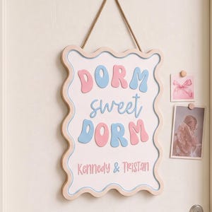 May include: A rectangular wall sign with a wavy border, featuring the text "DORM sweet DORM" in large, bubbly pink and blue letters, with the names "Kennedy & TRISTAN" printed in smaller pink text below, all suspended by a thin gold cord.