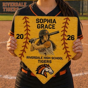 May include: A softball player holds a yellow home plate-shaped plaque with the name "SOPHIA GRACE" and a player illustration. The plaque also displays "RIVERDALE HIGH SCHOOL TIGERS" and the numbers "20" and "26". The player is wearing a black and orange uniform.