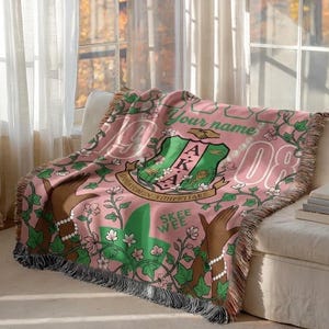 May include: Pink woven throw blanket with a green and white floral design, featuring the letters AKA and the number 08. The blanket has a fringe trim and is draped over a white sofa. The words "Your name" are also visible.
