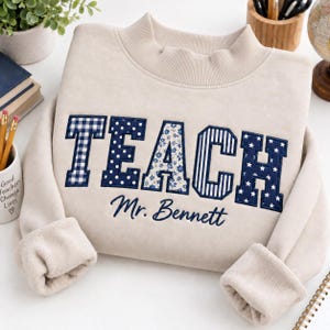 May include: A beige long-sleeve sweatshirt with a mock neck collar. The word TEACH is appliqued in large navy blue letters with varied patterns including gingham, polka dots, floral, stripes, and stars. Below, Mr. Bennett is embroidered in navy.