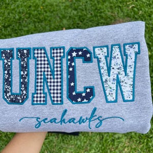 May include: A heather gray sweatshirt featuring the letters UNCW in large, fabric-appliqued block font. Each letter has a unique pattern: floral, black and white gingham, white stars on navy, and blue floral. The word seahawks is embroidered below.