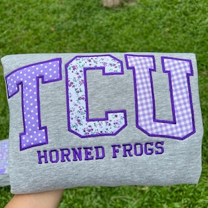 May include: A heather gray sweatshirt featuring the letters TCU in large, purple-bordered applique patches. The T has a purple polka dot pattern, the C has a floral print, and the U has a purple gingham check. The text HORNED FROGS is embroidered below.