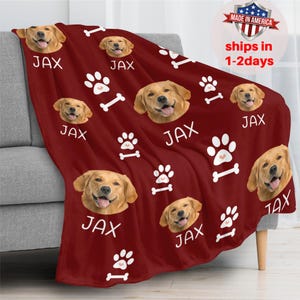 May include: A burgundy fleece blanket featuring multiple images of a golden retriever's face, paw prints, and bones. The name "JAX" is printed in white. The blanket is draped over a gray sofa.