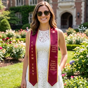 May include: A maroon graduation sash with gold trim and custom text. The sash has the words "CUSTOM TEXT" vertically printed on one side and space for a logo, photo, flag, or badge on the other. The sash is worn over a white dress.