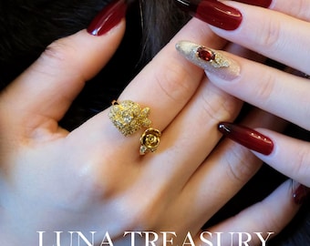 Può includere: Un anello d'oro con un design floreale, con piccole pietre e un dettaglio a forma di rosa. L'immagine mostra anche una mano con unghie lunghe e rosse e il testo "LUNA TREASURY" in bianco.