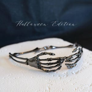 May include: A black skeleton hand headband with the text "Halloween Edition" in white script. The headband features two skeletal hands that appear to be holding the band together. The product is displayed on a white surface with a dark blue background.
