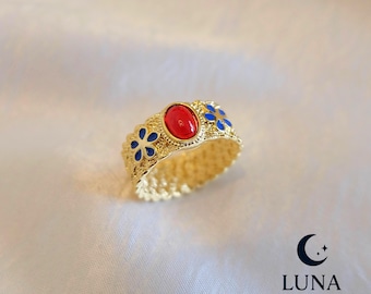 May include: Gold-toned ring featuring a central oval red stone and blue floral accents. The ring has a textured band and is set against a white background. The Luna Treasury logo is visible in the bottom right corner.